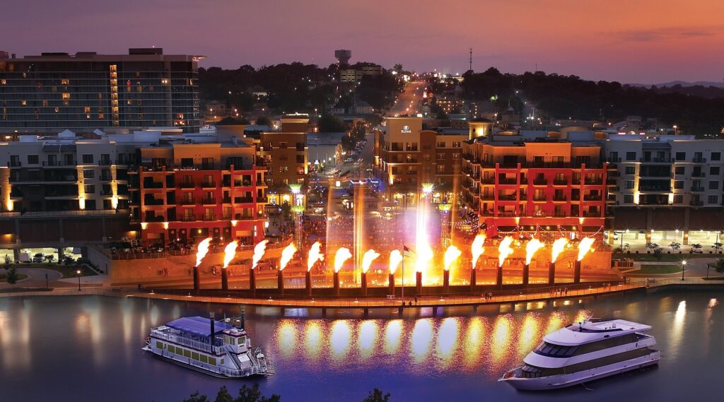 A serene cityscape at night featuring a river illuminated by lights and boats gently floating on the water's surface in Branson Landing