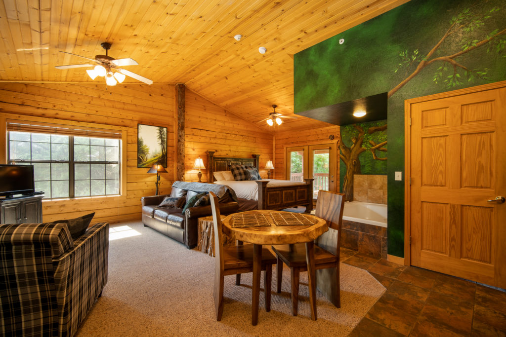 Interior of a Branson cabin with knotty pine walls and vaulted ceiling, a corner jacuzzi tub beside a painted forest mural, and a bed, sofa and dining table in one open room