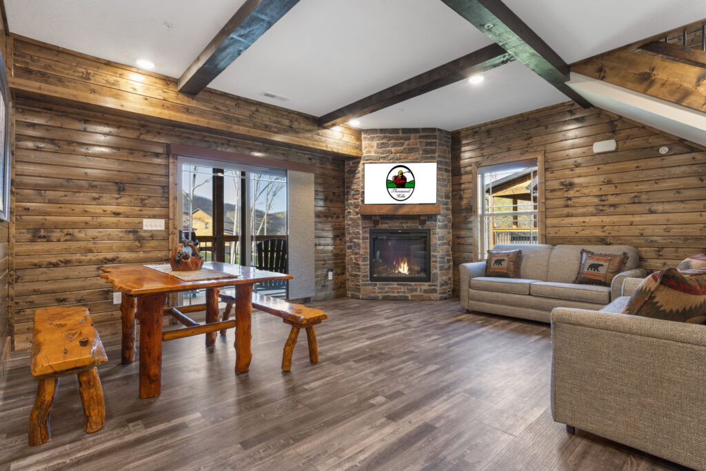Living room in a Branson cabin rental with a lit stone gas fireplace, exposed ceiling beams, knotty pine walls and a rustic log dining table