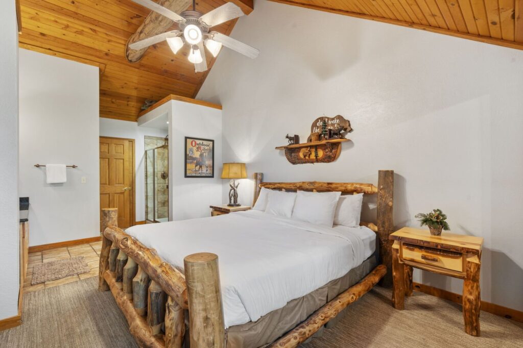 Bedroom in a Branson cabin rental with a log bed frame, vaulted pine ceiling, ceiling fan and an open door to the bathroom